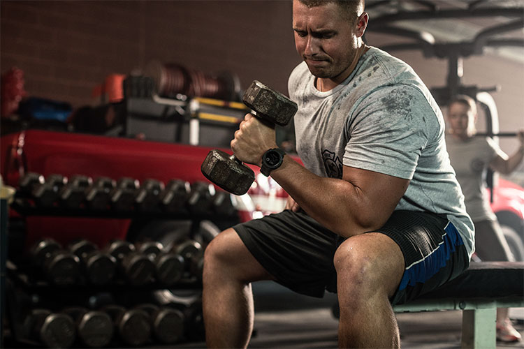 En man som tränar biceps på gymmet med en hantel i handen sittandes på en bänk. På handleden syns en svart sportklocka.